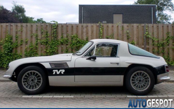 Early TVR Pictures - Page 81 - Classics - PistonHeads - The image features a classic car, painted in a striking combination of white and black, parked outside on a brick driveway. The car has a distinctive elongated nose and a sloping rear, characteristic of sports cars from the 1960s or 1970s. The car is positioned facing the left-hand side of the image, and the gear shift is clearly visible on the passenger side. The driveway is lined with a wooden fence that runs parallel to the car, and there are green plants growing on the fence, adding a touch of color to the scene. The sky in the background is overcast with clouds, suggesting a potentially cool or cloudy day.