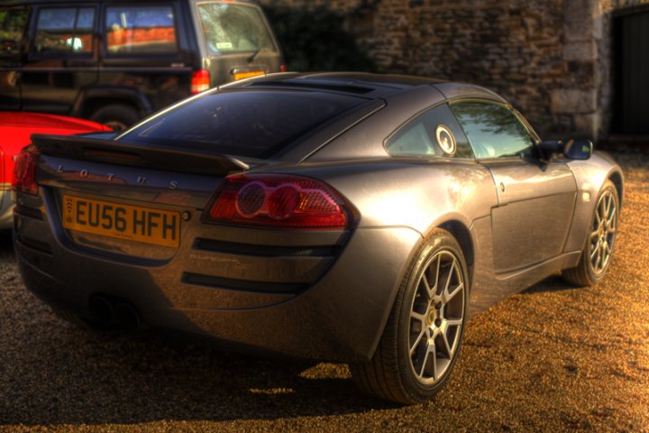 Pistonheads Issue Europa - The image showcases a very sporty and stylish grey two-door sports coupe parked on a gravel surface. The car is positioned at an angle that shows its rear side and features a distinctive taillight design with a set of European-spec model license plates. In the background, partially obscured, there's another vehicle parked on the edge of the lot. The scene conveys a relaxed, private parking area with no immediate sign of a main road or intensely populated area. The text "www.carfanatics.com" is visible in the lower right corner, suggesting the image may be from a car enthusiast website.