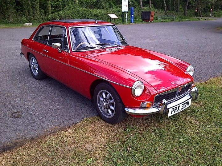 My mgb gt - Page 1 - MG - PistonHeads - The image shows a classic red sports car parked on a gravel driveway next to grass. The car has a prominent front grille, and its swept-back headlights and silver trim are characteristic of the era in which it was produced. The setting appears to be in a rural area with trees in the background. The vehicle's hood is open, allowing the engine to be seen. There are no visible license plates or distinctive markings that would identify the specific make or model.