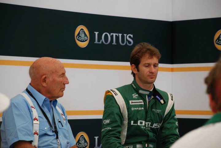 Pistonheads Goodwood - The image shows two men, one in a light blue shirt with a badge and the other in a vibrant green racing suit with logos, recognizable as a Lotus racing suit. They appear to be engaging in a conversation, possibly about a race or racing experience. The background suggests they may be in a setting related to Lotus racing, like a race track or event. The man in the green suit seems attentive and is looking towards the other man with a question on his face, suggesting an interview or a serious discussion about a technical or racing matter.