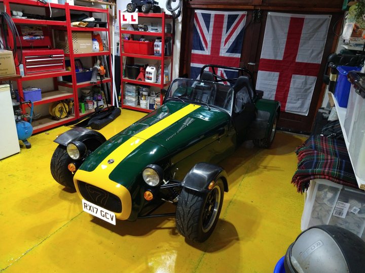 Not enough pictures on this forum - Page 73 - Caterham - PistonHeads UK - The image shows a small room with a yellow and green striped motorcycle prominently displayed. The floor is yellow, and there are red shelves on the wall behind the motorcycle. To the right of the photo, there's an open door revealing a white and blue striped flag hanging inside, suggesting a theme or country of origin. A small table with a few items on it is visible to the left side of the image. The room appears to be used for storage or display of vehicles.