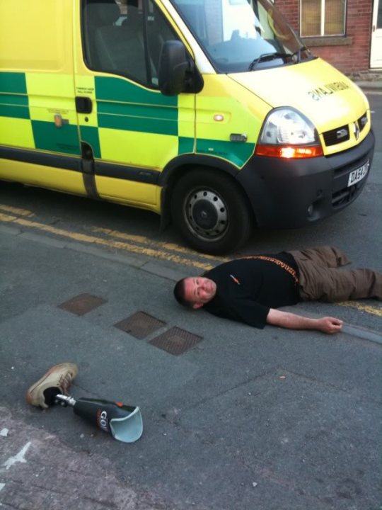 Ambiwlans Pistonheads Ambulance Dumbing - The image features a person lying on the ground next to a van. The van is green and yellow in color. The person appears to be tan and is clothed in brown pants and a dark shirt. The position of the person and the van suggests a context of being involved in an incident or accident. The van carries distinctive medical emergency signage, including red crosses and the number '245'. The medical cross signifies first aid help, while the number 245 typically indicates that medical personnel are available round the clock.