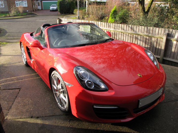 A close up of a car parked on a street - Pistonheads - This image showcases a vibrant red sports car parked in front of a brown wooden fence. The car, which appears to be a Porsche, is sleek and combines bright red and black color elements. Its shiny surface reflects the sky above and the reflection of the photographer's burgundy hoodie above it. The license plate of the car displays the year it was manufactured. The car's stance implies it is prepared to be moved shortly.