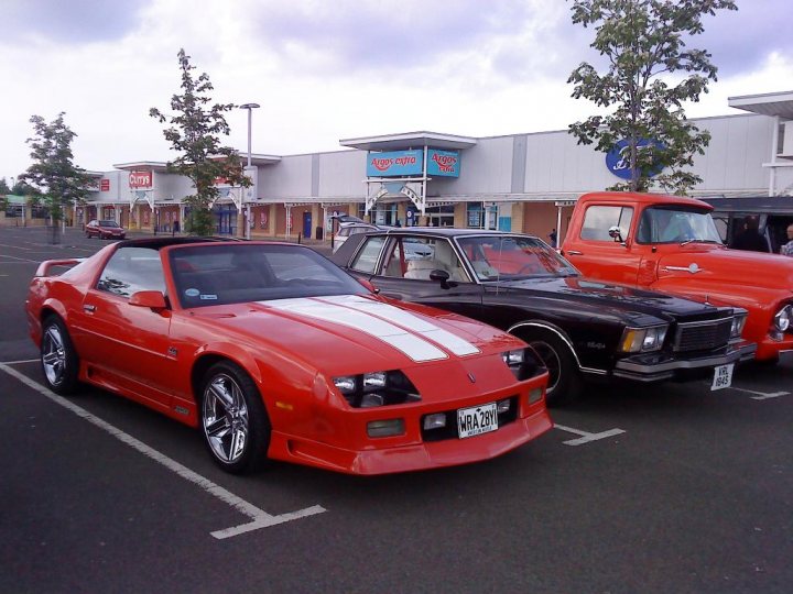 Cruisemeet Edinburgh Pistonheads - The image showcases a parking lot where two classic muscle cars are parked. The first car, a vibrant red with a white stripe on its hood, displays a sense of power and style. The second car, a sultry black with an orange driver's side door, adds a touch of mystery to the scene. These vehicles are parked next to each other, as if they are ready to roar into action at a moment's notice. Behind them, a building with a distinctive blue sign and a red sign contrasts with the minimalist architecture of the surrounding area.