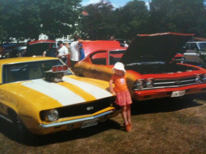 Our old Chevys - Page 1 - Yank Motors - PistonHeads - This image captures a scene from a car show with two vintage Pontiac GTO cars parked on a grassy field. One car is painted in vibrant shades of yellow and white, while the other one displays a bold combination of red and black. 

Among the attendees, a young girl stands next to the white and yellow car, her white hat and orange outfit adding a pop of color that complements the car's design. Her stance suggests she might be admiring the vehicle. 

In the background, a crowd of people can be seen, some of whom also appear to be observing the classic cars. They add a lively atmosphere to the event, contributing to the overall festive feel of the car show. 

The image gives an impression of a sunny day, as the sunlight casts a warm glow on the cars and their surroundings, enhancing the beauty of the vintage vehicles and the enthusiasm of the attendees.