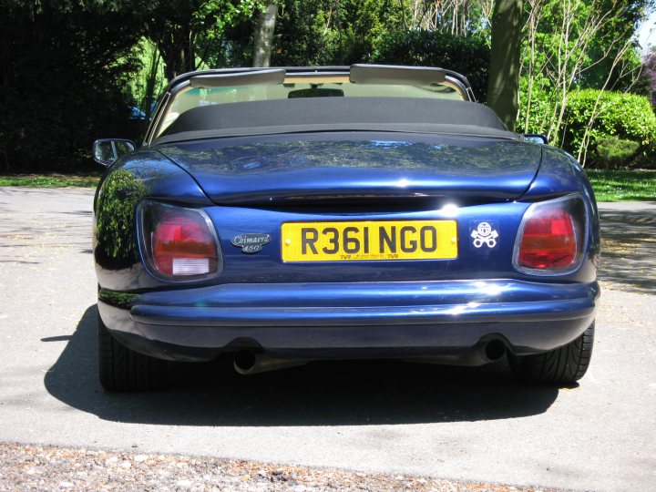 Where's your smiley? - Page 1 - Kent & Essex - PistonHeads - The image is a photograph of a blue R36 INO car positioned diagonally across a paved surface within what appears to be a park setting. The car has a visible UK-style license plate that spells out "R36INGO" on a yellow background. The vehicle's front end is visible, displaying a manufacturer's logo, round symmetric headlights, taillights with a red glow, and a partial view of a side mirror on the right side of the vehicle. The environment includes trees, grass and shrubs, with ample sunlight casting shadows on the ground and road. The style of the image is candid and appears to be taken casually without artistic composition, though it captures a unique moment in a specific location.