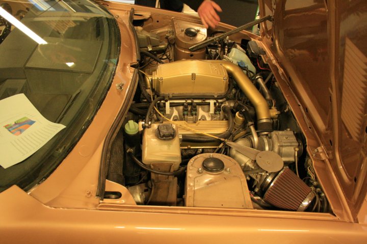 350hp from a 3.5 engine - Page 1 - Major Mods - PistonHeads - The image shows the open hood of a vintage auto, revealing a complex array of engine components. In the center of the visible assembly, there is a prominent golden fastener screwed into the trunk lid. The engine appears to be in disarray due to the mechanics of installation and dismantling. The hood is propped open with a long rod, held by a person partially visible on the left edge of the photo. It is a photograph characterized by the soft diffuse lighting of an interior setting, perhaps an auto maintenance shop.