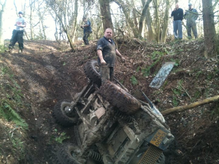 Pistonheads - The image depicts a hilly off-road terrain, where a person in a black top appears to be trying to extricate a tipped-over four-wheeled vehicle, which is coated in mud. The individual is gesturing towards the overturned vehicle with black tires and a metal frame, possibly indicating a problem or providing instructions. In the background, there are other people observing the situation, and the overall setting suggests a rough outdoor area with muddy ground.