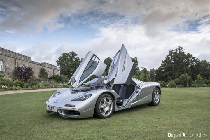 A black and white photo of a car parked in a field - Pistonheads - The image showcases a silver supercar parked on a manicured lawn in front of a stone building. The car's driver's side door and hood are open, revealing its sleek interior and engine. The sky is partly cloudy, suggesting a pleasant day, and the grass is lush and green, adding to the envisioned luxury. The overall setting speaks of a grand home or a salubrious retreat.