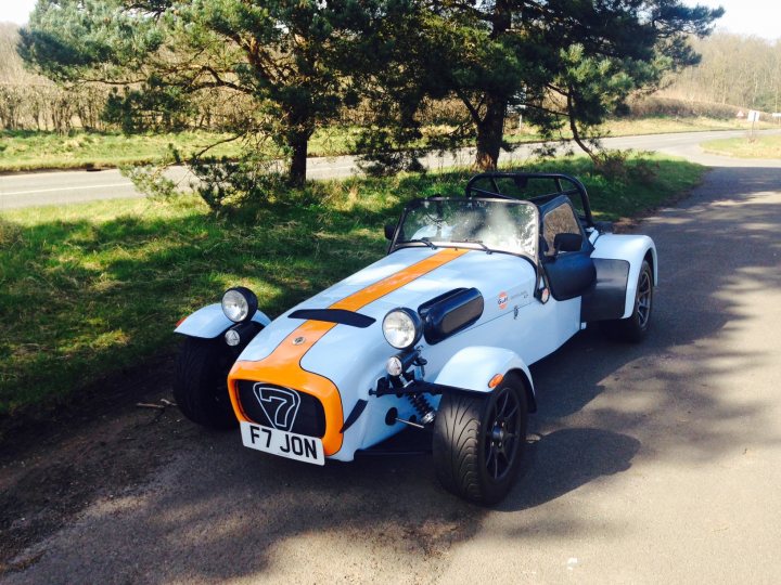 Show us your Caterham photos - Page 1 - Caterham - PistonHeads - The image shows a classic blue and white sports car parked next to a paved road. The vehicle is adorned with orange and white stripes and has the registration number F7 JON on the front. In the background, there are trees and the road curves to the right. The car appears to be in good condition, suggesting it's either well-maintained or new.