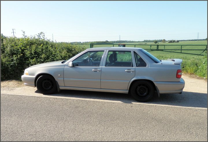 Steel wheels.Am I the only one? - Page 4 - General Gassing - PistonHeads - The image shows a silver Mercedes-Benz car parked on the side of a road. The car is parked next to a grassy area and appears to be in good condition, with recent use evident from the driver's side door being left open. The road itself is asphalt with white lines indicating lanes, and there's a somewhat blurred background suggesting a daytime outdoor setting with overcast or cloudy skies. In the top right corner, there's a wooden fence and some other shrubbery, reinforcing the rural or semi-rural nature of the location. The style of the car suggests it could be from the 1990s or early 2000s.