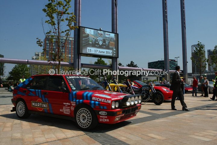 A car is parked on the side of the road - Pistonheads - The image captures a scene outside a public space, where a small gathering of people is taking place. The most striking feature is a Martini race car, painted in bold shades of blue and red, parked on a street lined with trees. The car is adorned with various stickers and a "Michelin" advertisement, indicating its association with the racing team. Other cars and motorcycles are also parked in the background, adding to the ambiance of a motor-related event or location. A few people are observing the parked vehicles from a distance, blending into the urban backdrop.