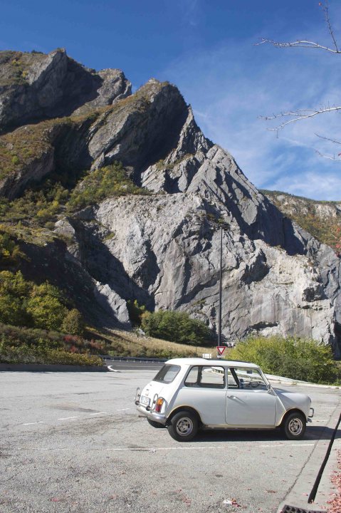 Returning after 38 years! - Page 1 - Classic Minis - PistonHeads - This image captures a serene scene in a deserted parking lot nestled at the foot of towering hills. A stone wall, part of the surrounding landscape's natural beauty, encloses the lot, adding a sense of tranquility to the image. A car is parked inside, all alone under the vast expanse of the blue sky. The hills, predominantly gray, rise majestically against this backdrop, their rugged texture contrasting with the car's smooth, pristine white exterior.