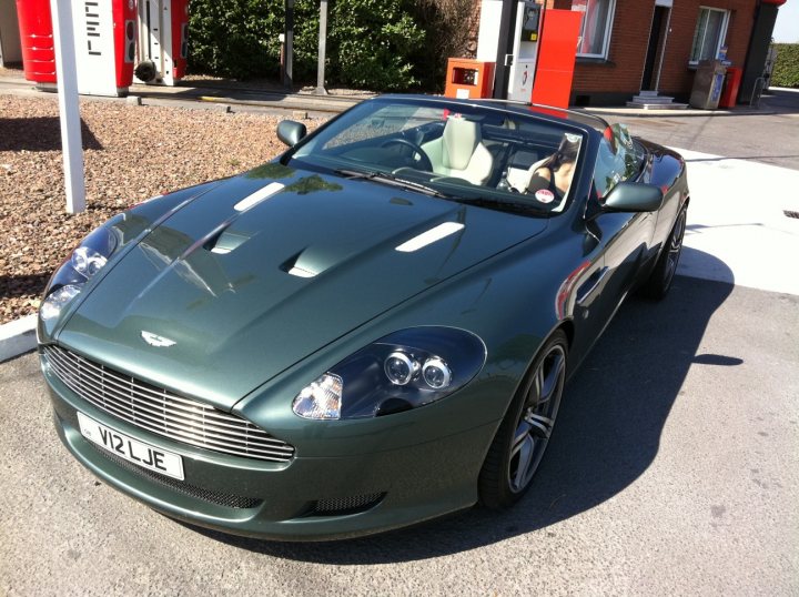 Pistonheads - This image features a sleek, modern green sports car parked on an asphalt surface outside a gas station. The car has a prominent windshield and is positioned in profile, allowing a clear view of the iconic round emblem on the front. The background is set against a natural landscape, with trees and shrubbery visible, adding to the serene outdoor setting. The photo appears to have been taken during the day.