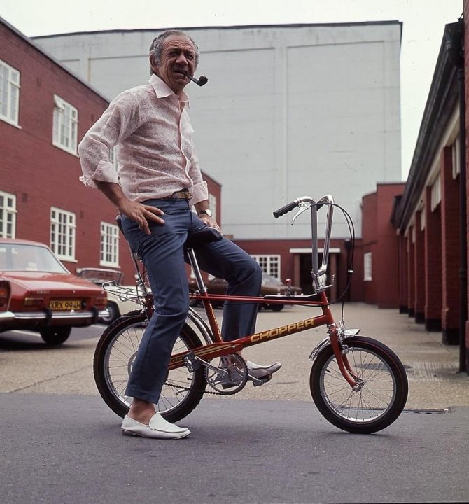 Pistonheads - The image is a vintage photograph, capturing a moment of a man with a mustache sitting on a red bicycle. He's wearing a shirt and pants, exuding an air of casual elegance. The setting appears to be an outdoor parking lot or a similar urban space, with multiple vehicles parked in the background.