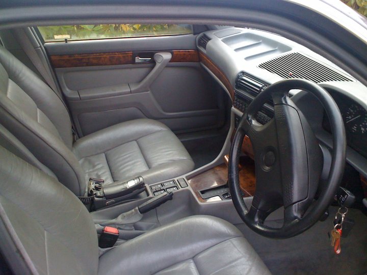 Pistonheads Shed Week - The image shows the interior of a car, focusing on the driver's seat. The car has a light beige interior color. The driver's seat has a modern design with a semi-circular dashboard and a central console visible in the driver's view. The steering wheel is black, likely with controls for the vehicle's features. The climate control buttons and the gear shift lever are in the center console. The door is open, and the armrest introduces a striking contrast with a red and black striped pad. The photo is taken from the perspective of the passenger seat, perhaps to present the layout and seating arrangement more clearly.