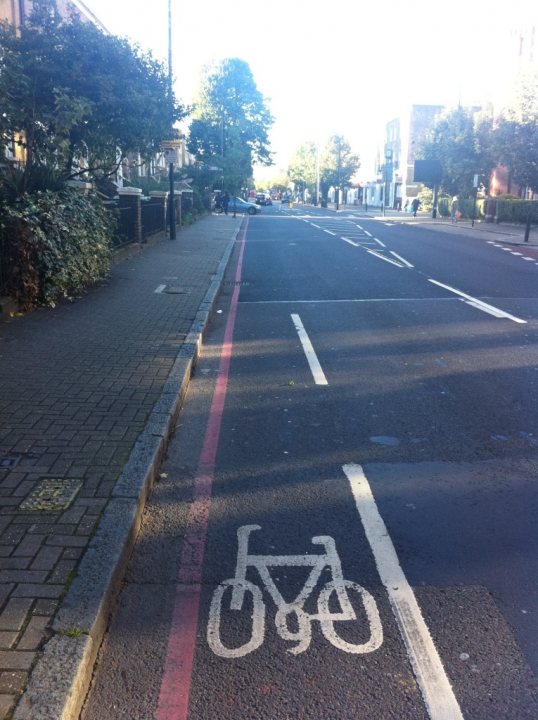 Pistonheads Bike Stand Knocked - The image shows an urban street scene viewed from the side of the road. A bicycle lane is clearly marked with white lines on the asphalt pavement. There is a striking pink line running along the edge of the pavement, perhaps for a different purpose or for aesthetic appeal. The buildings on the right side of the image give a sense of a densely populated area. A few cars are visible at the far end of the street, indicating light traffic. The sky is clear and blue, suggesting a sunny day.