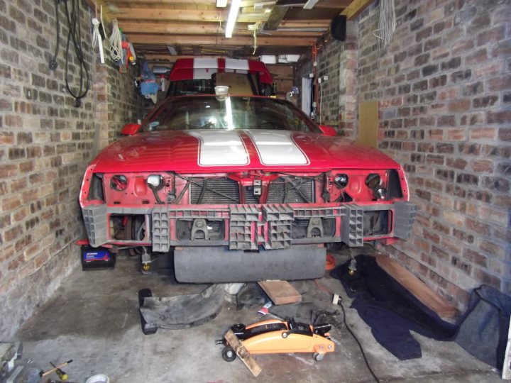 Who has the best Garage on Pistonheads???? - Page 56 - General Gassing - PistonHeads - The image shows a red and white race car parked inside a garage. The car appears to be a vintage model with a distinct front bumper design. The garage setting is crowded, with various mechanical parts and tools scattered around the floor, indicating that the car is currently undergoing modifications or repairs. The walls of the garage are made of brick, adding a rustic touch to the scene.