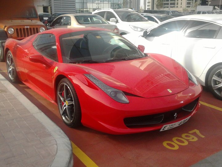 Oh to be the boss!! - Page 1 - Middle East - PistonHeads - The image features a vibrant red Ferrari parked in a parking lot alongside several other cars. The Ferrari, with its gleaming red body and striking yellow accents, is firmly positioned near a curb, which has the word "O097" painted on it. Its wheels and grill nicely accentuate the car's sporty design. A caution sign is visible in the background on the farthest right side of the image.