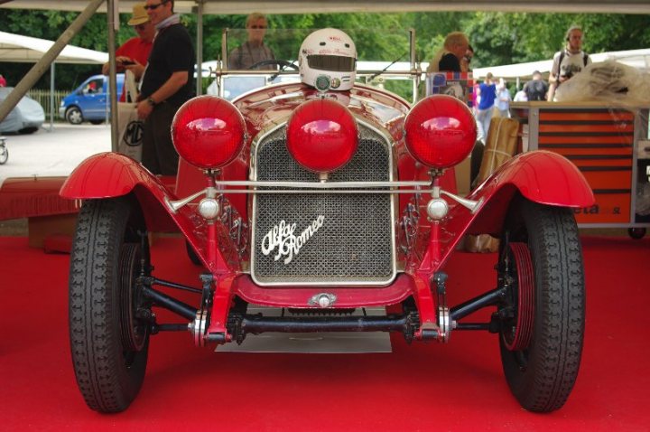 Anyone been to the Moving Motorshow before? - Page 1 - Goodwood Events - PistonHeads - The image depicts an antique red car positioned on a red carpet at what appears to be an auto show or event. The vehicle is a vintage model with noticeable features like a large grille, extra headlights in the front, and an aerodynamic design. The car is adorned with a round emblem in its center, possibly the brand logo. The backdrop of the image suggests a formal setting, with people standing in the background and a tented portion of what looks like a display area with other vehicles present. The presence of a person in the short front part of the car could imply that the vehicle might be on showcase.