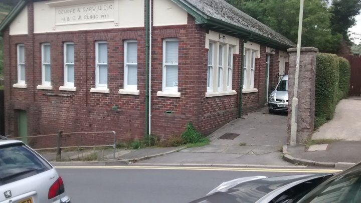 A car parked on the side of the road - Pistonheads - The image depicts a two-story brick building with a flat roof, featuring multiple windows. In the foreground, two cars are parked on the street; one vehicle has obstructed its rear window. The building has a sign indicating "Surgical Rathbone LTD." A metal gate is visible in front of the building, leading to a sidewalk and a smaller building in the background. There's also a compact car parked by a hedge, under the shade of a tree, in front of a brick fence.