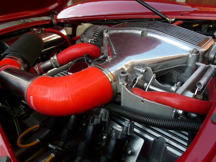 Megasquirt: true DIY? - Page 2 - Major Mods - PistonHeads - The image is a close-up photograph of the engine bay under the hood of a vehicle. The engine appears to be a V8 with a prominent red hose running along the engine surface. The shiny metallic surfaces of the engine components, along with the shiny black plastic of the air intake, reflect light, indicating that the car may be recently serviced or well-maintained. There is no visible text or distinctive logos in this section of the vehicle. The hose is attached near the air intake, which has a ribbed design with a rounded top. The focus is on the mechanical parts and the conduit, suggesting that the image may have been taken for an article about automotive engineering or engine maintenance.