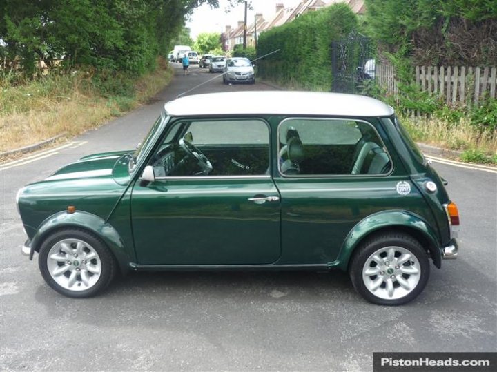 Buying An Older MINI? - Page 1 - Classic Minis - PistonHeads - The image depicts a small green car parked by the side of a road. The car has a hatchback design and features a compact size with a closed rear hatch. Its green exterior complements the silver five-spoke wheels which suggest it might be a keeper or a collector's item. In the background, we can see a person walking, although the focus of the image is clearly on the car. The setting is a residential area with a tree-lined street and a fence visible in the distance.