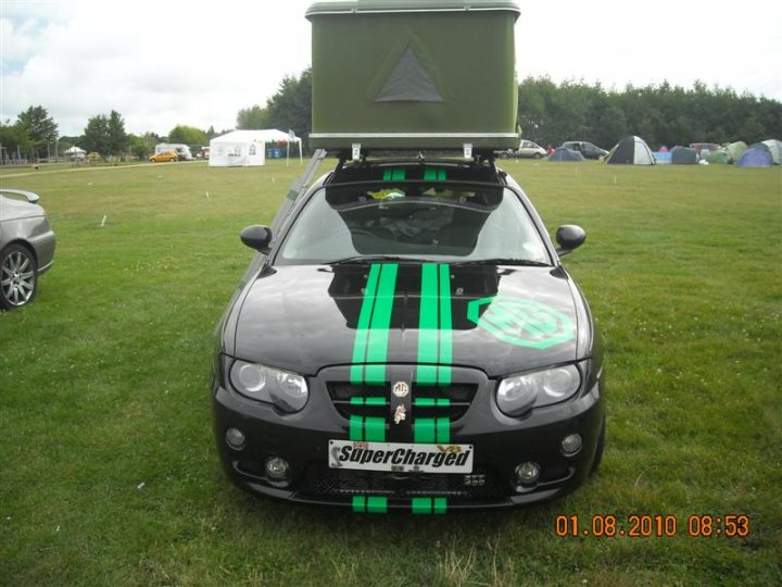 Pistonheads - In the heart of a grassy field, a black car with a striped roof leads a convoy of camping trailers in what appears to be a camping trip. The car stands out with its unique roof and a charging bay attached to it, hinting at its purpose in this setup. Tents of various shapes and sizes are set up around the arc of parking, creating a cozy camp. The image captures a moment of anticipation, as if the journey is just about to start or has just ended. The date '01.08.2010 08:53' and the car's registration 'SUPERCHARGED' hint at memories of a trip taken in August 2010.