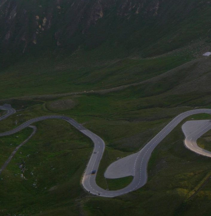 2 young guys killed in Austrian Alps racing. Came off a clif - Page 22 - General Gassing - PistonHeads - This is an aerial photograph of a winding mountain road with a sharp, sudden curve from the viewer's perspective. The road curves gently before this point, and there is a fork in the road where one path continues straight and the other bends to the left. The terrain appears to be rugged and mountainous, with a variety of green tones suggesting an abundant landscape. The overall color palette is muted with greens from the landscape and a touch of blue in the sky. The vehicles in the road are tiny, indicating the scale of the landscape.