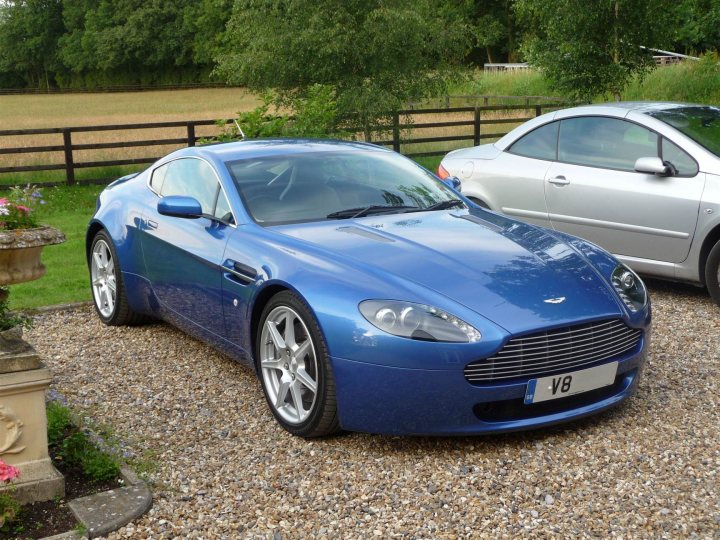 Finally took the plunge!  *happy bunny* - Page 1 - Aston Martin - PistonHeads - The image shows a large, blue, limited edition sports car parked on a rocky surface next to a silver car. The blue car has a shiny finish and sleek, aerodynamic design typical of luxury models. It features custom rims and a license plate with the letters "V8," indicating it may be a special edition or part of a collection. The car is situated in an outdoor setting, likely a driveway or garage, with greenery and grass in the background. The overall scene suggests a well-maintained garage or storage area for high-end vehicles.