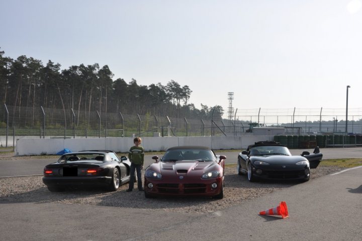 Clark Hockenheimring Jim Pistonheads - This image captures a scene of a young man standing amidst a variety of exotic and classic cars in an open-air parking lot or race track. There are three cars lined up in the foreground: a black convertible, a red classic car, and a black sports car. In the background, there are trees and a fence lining a race track or off-road area. The sky is overcast, and there is an orange traffic cone on the ground next to the red car, possibly indicating a recent activity or a forthcoming race.