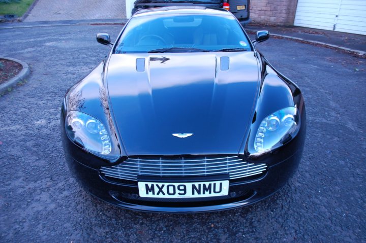 Pistonheads - The image depicts a black Aston Martin DB9 car parked in a driveway during the day. The car's body reflects the sunlight, highlighting its sleek lines. A key fob hangs from the car's side mirror, and a white license plate is present at the front. The scene suggests a residential area.