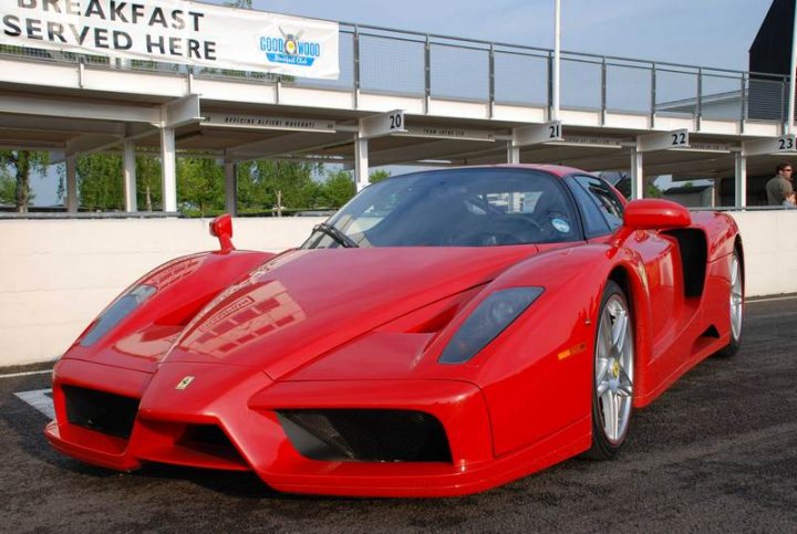 Pistonheads Supercar Sunday - This image captures a vibrant scene on a racetrack. Dominating the frame is a striking red Ferrari sports car, parked prominently in the foreground. The car's sleek design and the gleam of its shiny shell reflect the glaring sunlight. 

The vehicle is parked in a garage area of the circuit that has a brick wall in the background. The signage suggests a race environment. There are also a few blurred spectators visible in the distance, adding to the ambiance of the racing event. 

The overall colors in the picture are warm, with the automobile's red hues contrasting sharply with the gray and white of the garage and the spectators. The presence of the Ferrari, a symbol of speed and luxury, instantly conveys the thrill and excitement of high-performance automobilism.