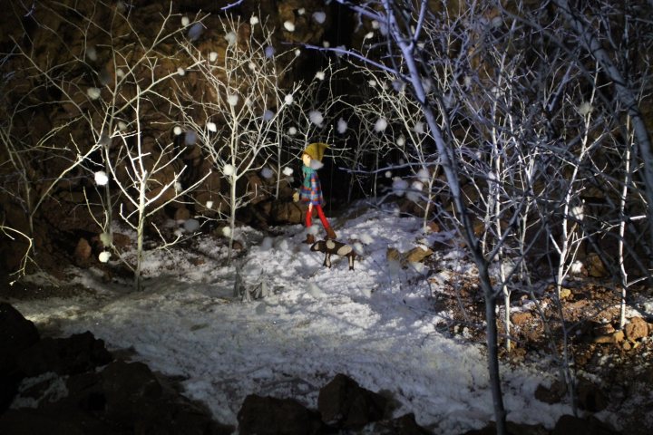 Santa in a cave  - Page 1 - South West - PistonHeads - This image captures a serene winter night scene in a forest. There is a vignette of a snowy pathway filled with snow-laden bare trees and large rocks. In the middle of the forest, there's a small wooden fence that adds a rustic touch to the scene. Amidst this wintry landscape, a small figurine of a girl wearing blue and red is placed, as if watching over the enchanting forest. This picture has an enchanting atmosphere enhanced by the falling snowflakes illuminated by the soft fairy lights scattered throughout the forest, creating a mystical aura.