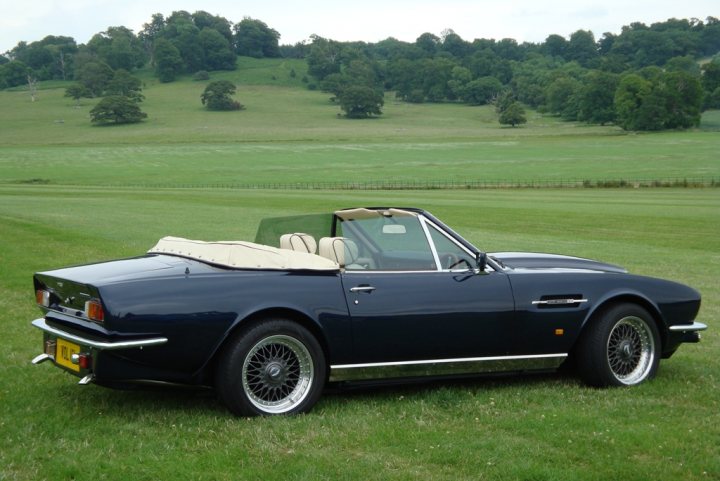 Pistonheads - The image depicts an open-top, blue convertible car parked in a grassy field. The car is styled in an old-fashioned manner, reminiscent of classic roadsters. It features four-cylinder wire wheels and a chrome exhaust pipe. In the background, there is a line of trees visible in the distance. The sky is overcast, suggesting it might be a cool or cloudy day. The setting appears to be peaceful, with no other vehicles or people in sight.