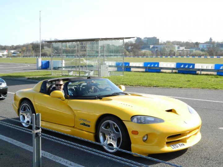 Pistonheads - The image portrays a vibrant scene on a parking lot. At the center of the image, a yellow sports car is parked, sleek and shiny. Two individuals are enjoying a road trip in this sports car, as evidenced by the two open windows, inviting the outside air in. They are seen driving the car, their attention focused on the road ahead. The background of the photo features an airport runway, possibly indicating their location or direction of travel. The day is bright and sunny, casting a warm glow on the entire scene.