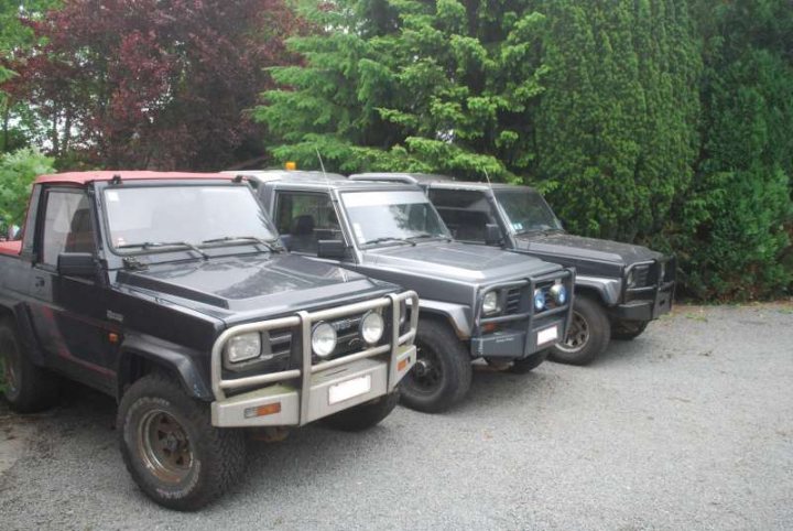 The Mud Squad - Page 1 - Readers' Cars - PistonHeads - The image shows three parked vehicles, which look like off-road or utility types, in a gravel lot with trees in the background. The vehicles are lined up, occupying different positions from left to right in the frame. Two of the vehicles are angular and appear to have black and silver color schemes, while the third vehicle is a mix of red and black and has a more rounded, jeep-like design. The lighting suggests it might be daytime.