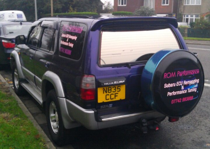 Pistonheads - The image shows a black SUV parked on a street. The car has a spare tire mounted on the back door, which is slightly ajar. On the tailgate of the car, there are bright stickers with contact information and a company logo, along with the text "ROM Performance SUBARU Ecu Re-Mapping & Performance Tuning". The car seems to be parked next to another vehicle, and there are houses visible in the background.