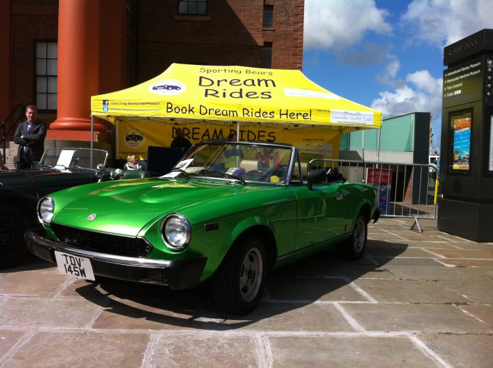 Lets see a picture of your classic(s) - Page 278 - Classic Cars and Yesterday's Heroes - PistonHeads - In the image, a green sports car is parked on a brick walkway. The car is facing towards the left of the frame and is partially shaded by a large yellow tent.  The tent has the words "Dream Rides" and "Book Dream Rides" on it, suggesting that it's a part of an event or promotion.  There are people milling around in the background, adding a sense of activity to the scene.  In the far right, a bus is visible, possibly indicating a public transportation route nearby.  The overall setting appears to be a public outdoor space, possibly on a city street.