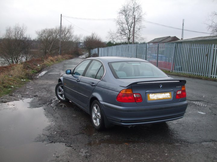 Pistonheads - The image captures a scene of an old gray BMW parked on a street. The car is slightly tilted to one side, parked facing the camera. The street is lined with lush trees, and a fence is visible in the background. Interestingly, the puddle under the car reflects just one leg of the car, adding an unusual and intriguing element to the photograph. This close-up image offers a detailed view of the car, emphasizing its age and condition.