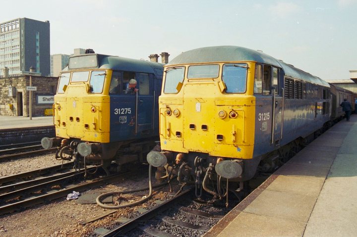 Loco sheds and other railway buildings.... - Page 16 - Boats, Planes & Trains - PistonHeads UK - The image captures a serene scene on railway tracks. Dominating the frame are two striking yellow trains, their vibrant color contrasting with the darker hue of the steel track they're parked on. They are stationary, awaiting their next journey, suggesting a moment of stillness in an otherwise busy transport network.

The backdrop is a quiet street corner, adding to the tranquility of the scene. A building can be seen in the distance, perhaps indicating a small town or city area near the tracks. The sky above is overcast, casting a soft light that seems to highlight the trains' yellow color even more.

The photo appears to be an old photograph, as suggested by its grainy texture and aged appearance. This gives it a sense of nostalgia, evoking memories of times gone by when train travel was more prevalent. Despite being inanimate objects, one can almost hear the chugging of the trains and feel the vibration of their engines underfoot.