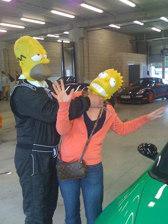 Random photos - Page 537 - Photography & Video - PistonHeads - The image is a humorous studio-like photograph, where two people are donning oversized costumes, imitating the characters Bart Simpson and Lisa Simpson from the animated television series "The Simpsons". They are playfully confronting each other near some cars. The person dressed as Bart, located on the left, and the person dressed as Lisa, centered in the frame, are the main subjects of the photo, and their exaggerated expressions and costumes add a comic element to the scene. The background shows typical cars and a stadium setting, but the focus remains on the two individuals in costume. There are no visible texts in the image.