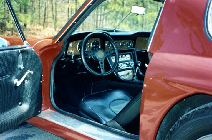 Grantura Griffith Vixen Tuscan Dash Pictures - Page 1 - Classics - PistonHeads - This image shows the interior of a classic car, revealing a distinctive orange and black color scheme. The perspective is from the side, allowing a clear view into the vehicle. The car's cockpit is visible, featuring a steering wheel and various gauges and controls that are typical of old automobiles. The driver's side door is open, suggesting that no one is presently. The car is parked, and the outside environment suggests a forest or wooded area.