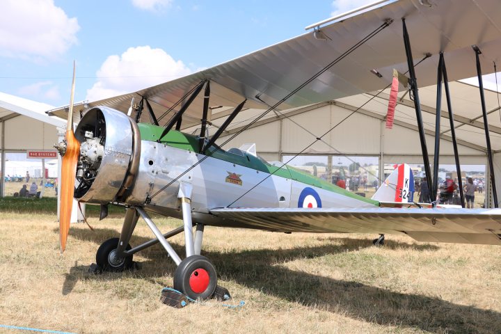 RIAT 2018 - Page 8 - Boats, Planes & Trains - PistonHeads - The image showcases an open-air airplane hangar, with the main focus being an old-fashioned propeller plane. The plane, painted in a white and green livery, has a propeller and a spinner on the nose. Its wings are adorned with a design featuring red, blue, and white stripes, and the tail has a small Union Jack symbol. The plane is stationed on the ground, with its wheels touching the soil. The hangar's structure is visible in the background, with support poles stretching across the space. The setting seems to be in an outdoor field, possibly during an airshow or exhibition, as evidenced by the partially visible spectators.