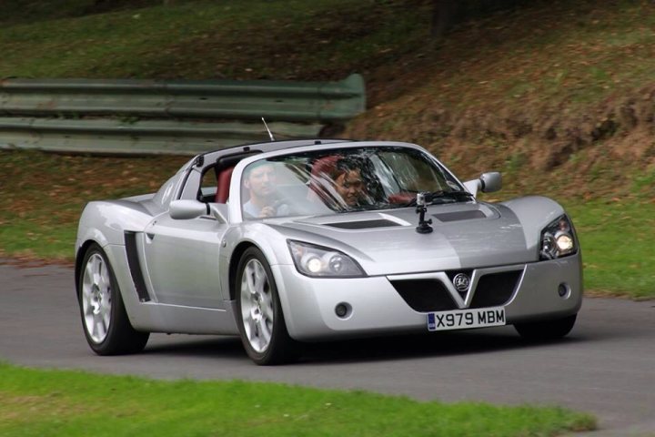 Prescott Revival - Sunday 10th August 2014 - Page 1 - South West - PistonHeads - The image captures a vibrant outdoor scene where a man is driving a silver sports car on a winding road. The driver, whose face is visible, is intently focused on the road ahead. The car is embellished with a sleek black convertible top and is equipped with a horn. The backdrop of the image provides a contrast to the speed and engineering of the car, as it showcases a serene landscape filled with lush greenery and gentle hills.