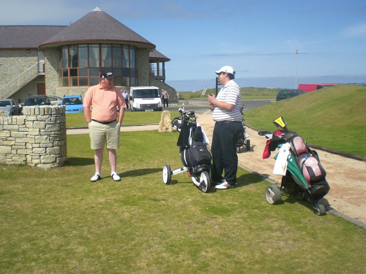 A man and a woman standing next to a motorcycle - The image captures a serene scene on a cloudy day. Two golfers are seen on a lush green golf course with a clubhouse in the background. One golfer, holding a bag of clubs, has finished or is about to start a round, while the other is approaching, presumably ready to play. Their golf bags are open, indicating that they are either preparing for or returning from a game. The presence of cars in the background suggests that this location is accessible by road.