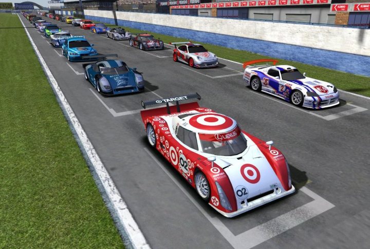 Mods Sims Tracks Driving Pistonheads Find - The image showcases a thrilling scene from a racing game. The track curves gently with a grassy embankment and a solid retaining wall. The cars are emblazoned with advertisements, prominently featuring a Target logo, indicating they could be modeled after the well-known Target Car. The vehicles are diverse, exhibiting different makes like Mercedes, Cadillac, and several more, each with its unique design and color schemes. The red, white, and black color scheme of one car in particular stands out starkly against the green track. The track is devoid of any obstacles or debris, suggesting that the race is currently in progress or just about to begin.