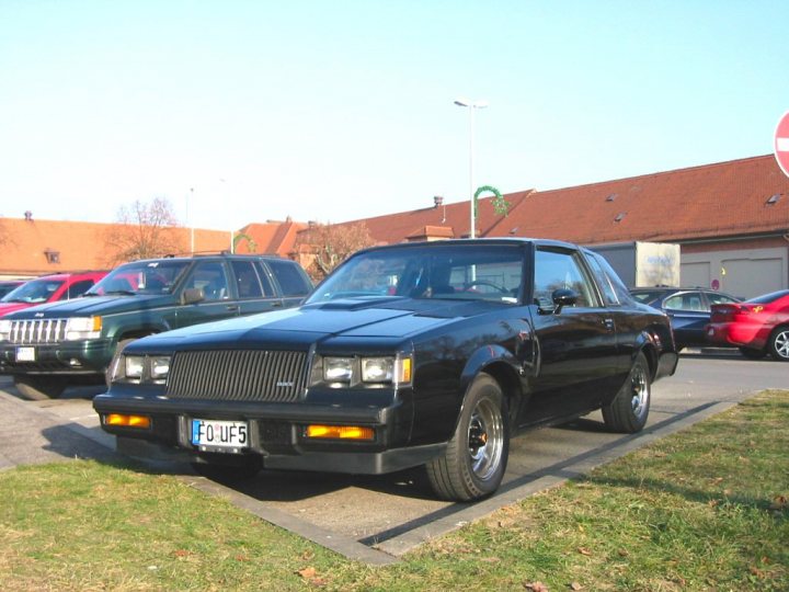 1987 GNX - Page 1 - Readers' Cars - PistonHeads - The image features a black coupe parked in a parking lot during the daytime. The car is positioned at an angle, showcasing one of its sides. The parking lot appears to be a public space as it has public parking spaces marked by white lines, and a "no exit" sign is visible in the background.