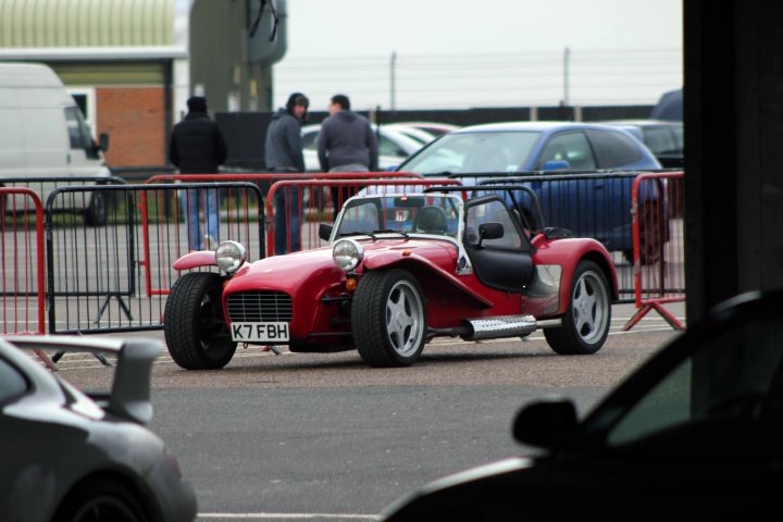 Track day at Snetterton Friday 15th February - Page 1 - East Anglia - PistonHeads - The image is set in an open street area, featuring a red, sports car with a convertible top currently down. This car is parked, and in the background, there are several other vehicles in a parking area, enclosed by a fence. A few pedestrians are standing at a distance, seemingly unrelated to the parked cars. A large building can be seen in the background, indicating the possibility of a commercial or industrial setting. The scene suggests it might be in an urban environment, as there are multiple types of vehicles present.