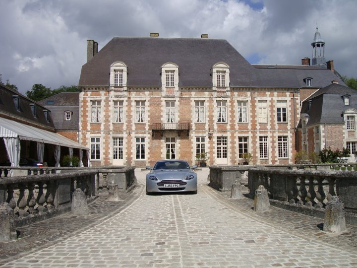 French trip - Page 1 - Aston Martin - PistonHeads - The image showcases a three-story mansion with a distinctive grey roof. The mansion features numerous windows and a prominent arched entrance. A black sports car sits parked in front of the house, adding a touch of luxury to the scene. The entry to the house is a long, wide driveway that stretches down to a quaint roadway adorned with a low stone wall and traditional European cobblestones.